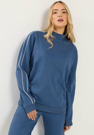 Blonde vrouw die een blauw lang mouw shirt en bijpassende broek draagt met witte strepen langs de mouwen en zijkanten van de broek, staand tegen een lichte muur.