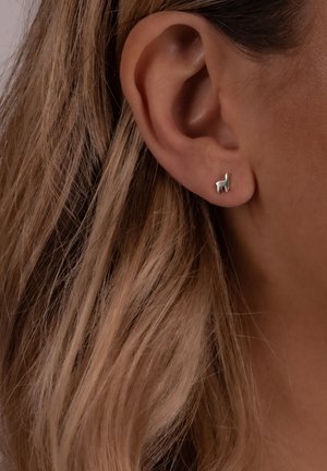 Sudraba lamu formas stud auskars iestiprināts ausī, ar gludu tekstūru un vienkāršu dizainu fona viļņainajiem gaiši brūnajiem matiem.