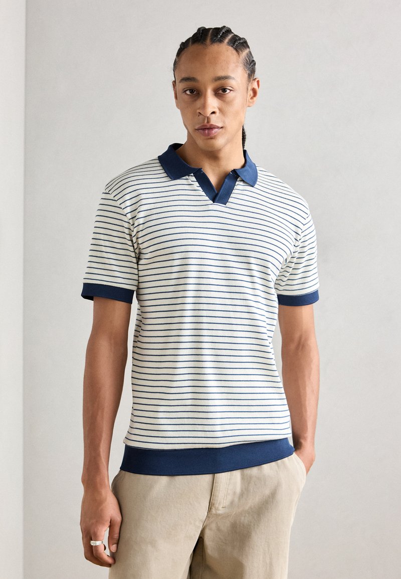 Polo rayé en blanc et marine, avec un col et des poignets côtelés. Tissu doux, à manches courtes et à coupe ajustée.