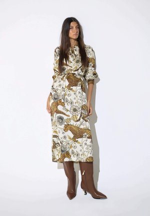 Mujer con cabello largo y oscuro que lleva un vestido blanco con grandes estampados de leopardo y flores, combinado con botas marrones de tacón alto, de pie contra una pared blanca.