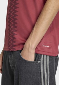 Röd funktionsskjorta med texturerat tyg, prydd med ett zigzag-mönster på ena sidan och texten "CLIMACOOL" nära fållen. Kombinerad med mörka jeansshorts.
