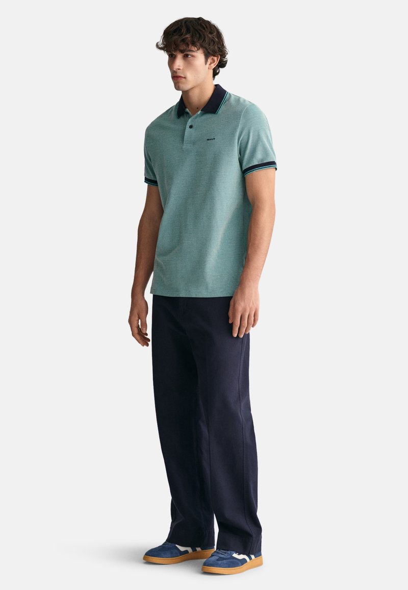 Hellgrünes Poloshirt mit einem marineblauen Kragen und Details, kombiniert mit dunkelblauen Hosen und blauen Sneakers mit weißen Akzenten.