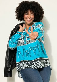 Blauwe blouse met patroon met abstracte ontwerpen en de tekst "HAPPY". Zwarte leren jas over de schouder gedrapeerd, gecombineerd met donkere spijkerbroek.
