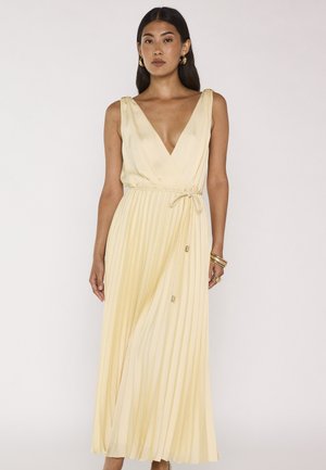 Donna che indossa un vestito plissettato giallo pallido senza maniche con scollo a V profondo e cintura annodata in vita, accessoriata con orecchini a cerchio dorati e braccialetti.