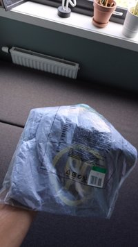 Priehľadné plastové balenie obsahujúce zložený šedý odev s jemným logom a tlačenými pokynmi na údržbu na zadnej strane.