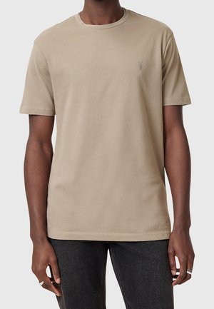 Uomo che indossa una t-shirt beige a maniche corte con scollo a girocollo e un piccolo logo ricamato sul petto sinistro e jeans neri.