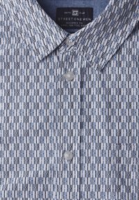 Košilové tričko s krátkým rukávem v geometrickém modro-bílém vzoru, s límečkem na knoflíky a průhledným plastovým knoflíkem vpředu.