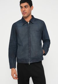 Τζιν μπουφάν με γιακά, σκουρό μπλε χρώμα, φερμουάρ μπροστά και υφασμένο σχέδιο. Διαθέτει πλαϊνές τσέπες και μακριά μανίκια.