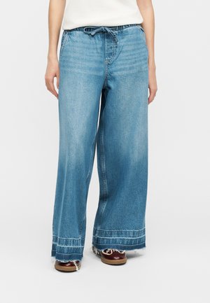 Persoană purtând blugi din denim albastru cu picioare largi, talie cu șnur și margini zdrențuite, asortați cu pantofi maro pe un fundal alb.