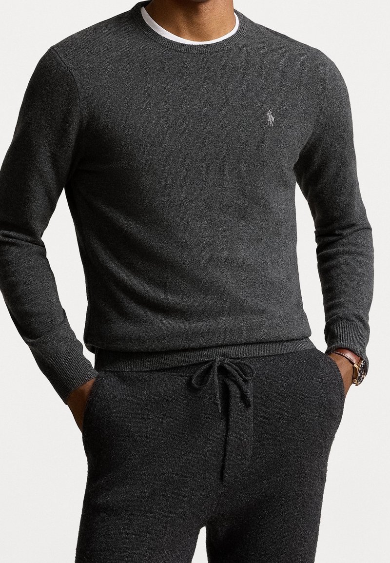Homme portant un pull gris foncé à manches longues et un pantalon de jogging assorti, mains dans les poches et une montre au poignet gauche.