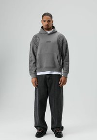 Grijze hoodie met patroon op de mouwen, een voorzak en tekst. Draagt over een wit T-shirt en gecombineerd met donker baggy jeans en zwarte schoenen.