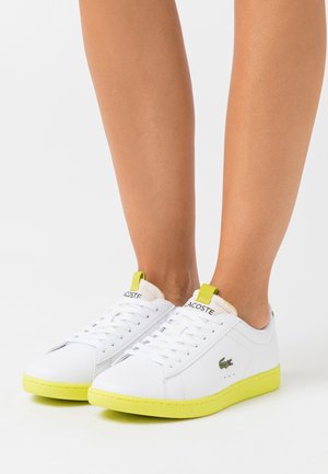 Vita sneakers i mjukt lädermaterial och med texturerad yta. Har klara gula sulor och detaljer, med en liten grön logotyp på sidan.