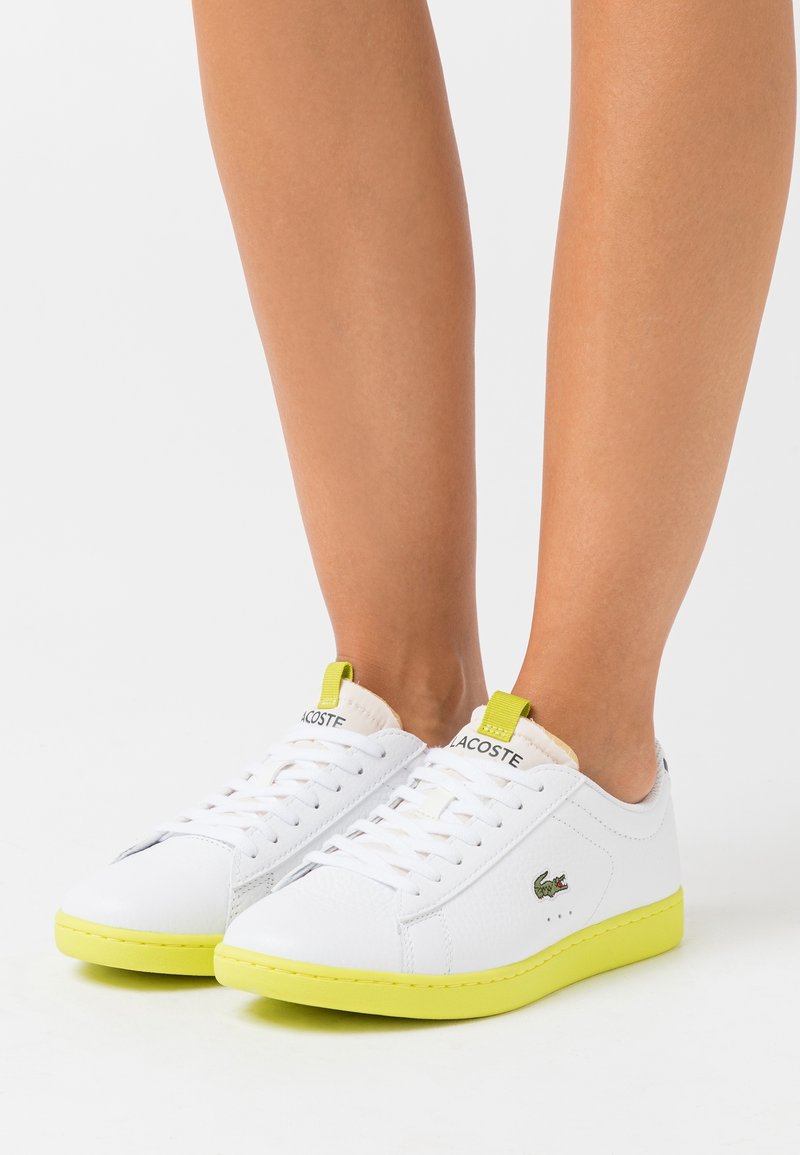 Vita sneakers i mjukt lädermaterial och med texturerad yta. Har klara gula sulor och detaljer, med en liten grön logotyp på sidan.