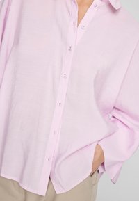 Lys rosa knapp-up skjorte med krage, lange ermer og bølget hem. Stoffet virker mykt og litt teksturert. Har diskrete knapper.