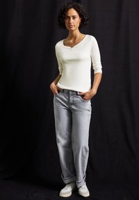 Vit långärmad tajt topp med rundad halsringning, i kombination med ljusgrå raka jeans och vita sneakers, mot en mörk bakgrund.