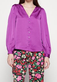 Blusa in raso viola con scollatura a V, maniche lunghe a sbuffo e chiusura con bottoni sulla parte anteriore. Abbinata a pantaloni neri con motivo floreale.