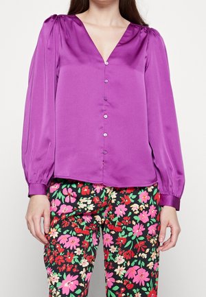 Lila Satinbluse mit V-Ausschnitt, langen Puffärmeln und Knopfverschluss vorne. Kombiniert mit schwarzen, floral gemusterten Hosen.