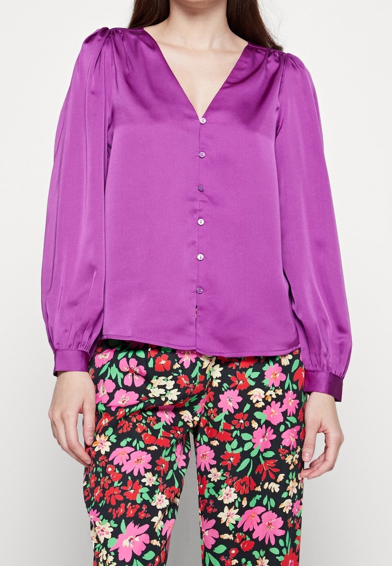 Blusa in raso viola con scollatura a V, maniche lunghe a sbuffo e chiusura con bottoni sulla parte anteriore. Abbinata a pantaloni neri con motivo floreale.