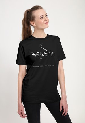 Ung kvinde med langt hår sat op i hestehale iført sort T-shirt med teksten "GENESIS" og "FOLLOW YOU FOLLOW ME", kigger op og smiler.