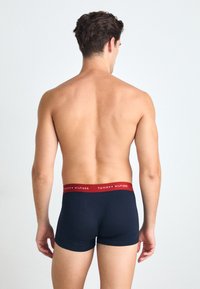 Sötétkék boxeralsó piros elasztikus derékrésszel, rajta a "TOMMY HILFIGER" logóval, hátulról nézve, világos háttér előtt.