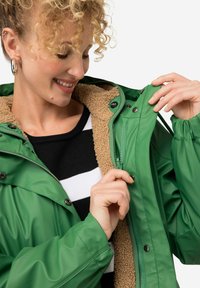 Giacca impermeabile verde con una fodera in fleece beige, dotata di bottoni a pressione e chiusura con zip. Camicia a righe bianca e nera visibile sotto.