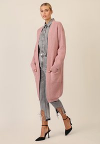 Lange, roze gebreide cardigan met ribbels, open voorkant en twee zakken, gecombineerd met grijze spijkerbroek met franjes en zwarte hakken.
