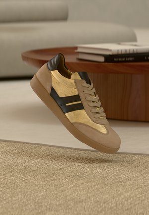 Beige and gold sneaker with black stripes hovering above a tan woven carpet, near a round wooden table with stacked books.