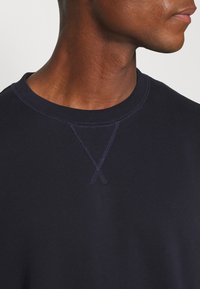 Gros plan d'une personne portant un sweat-shirt à col rond bleu marine avec un détail de couture visible au niveau du col.