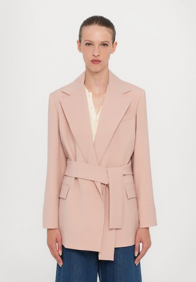 Blazer rosa chiaro con cintura, ampie rever e due tasche frontali. Il tessuto sembra liscio, abbinato a una camicia color crema sotto.