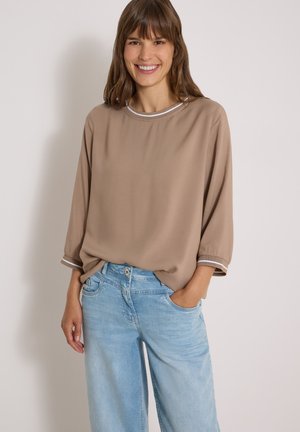 Femme souriante aux cheveux bruns portant un haut beige ample avec des rayures blanches sur les manches et un jean bleu clair, debout, la main dans la poche.