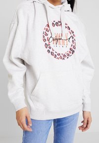 Osoba v světle šedé mikině s kapucí s růžovým leopardím potiskem v kruhu a nápisem „Tommy Jeans“, v kombinaci s modrými džínami, stojící s uvolněnými rukama.