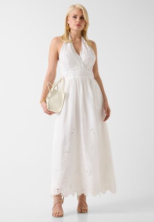 Donna bionda in piedi, indossa un abito lungo bianco ricamato senza maniche, tiene una borsa beige e indossa orecchini dorati e sandali.