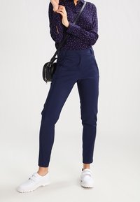 Granatowe eleganckie spodnie o slim fit z kieszeniami z przodu, w połączeniu z ciemną wzorzystą bluzką i białymi sneakersami. Czarna torebka na ramię.