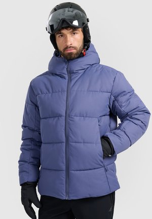 Mann trägt eine blaue Steppjacke, schwarze Skihandschuhe und einen schwarzen Skihelm mit reflektierendem Visier, steht vor hellgrauem Hintergrund.