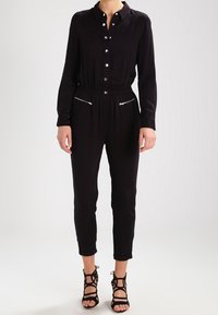 Une combinaison noire à manches longues, avec un col, une fermeture à boutons, une taille élastique et des poches zippées. Associée à des sandales noires à lanières.