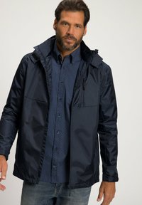Navy wasserfeste Jacke mit Reißverschluss vorne, verstellbarer Kapuze und Brusttasche. Kombiniert mit einem gemusterten Hemd mit Knöpfen und Jeans.