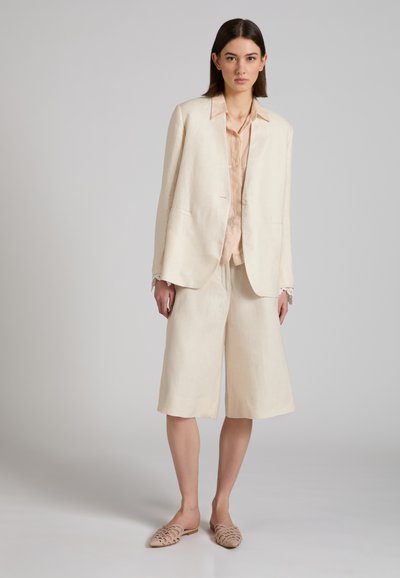 Vrouw die een beige oversized blazer draagt, bijpassende wijde korte broek tot op de knie, lichtroze blouse en beige geweven platte schoenen, staand tegen een effen achtergrond.