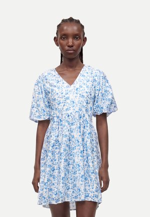 Donna con capelli intrecciati che indossa un vestito bianco con motivo floreale blu, scollo a V e maniche corte a sbuffo, in piedi davanti a uno sfondo chiaro.