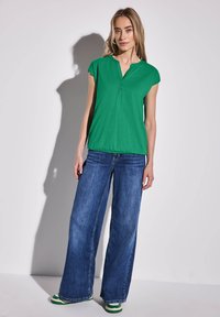 Zelená košile s krátkým rukávem a knoflíky, s utaženým lemem, spárovaná s volnými modrými džíny a zeleně pruhovanými botami na světlém pozadí.