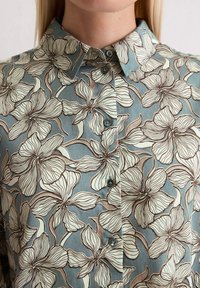 Floral button-up shirt in teal, featuring large white and brown flower patterns, a pointed collar, and two dark buttons on the front.