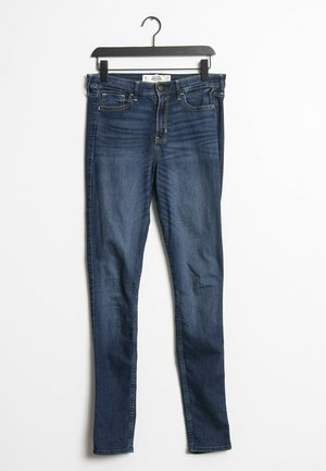 Dark blue denim skinny jeans with a mid-rise waist, five pockets, and slight fading detail. Features a zipper fly and a narrow leg opening.