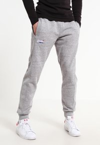 Graue Jogginghosen aus Baumwolle mit schlanker Passform, ausgestattet mit einem elastischen Bund, Seitentaschen und einem kleinen Logo am Bein. Kombiniert mit weißen Turnschuhen.