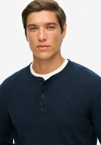 Jonge man met kort bruin haar, die een donkerblauwe henley shirt met drie knopen over een witte ronde hals top draagt.