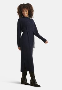 Abito in maglia blu navy con trama a coste, maniche lunghe e vita con cintura; presenta una spacco laterale ed è abbinato a stivali neri al ginocchio.