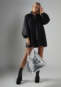 Camicia nera oversize con fiocco, abbinata a una borsa hobo in metallo argentato. Il modello indossa tacchi a piattaforma neri e calze grigie.