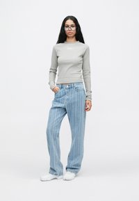 Νεαρή γυναίκα με μακριά σκούρα μαλλιά, φορώντας στρογγυλά γυαλιά, ανοιχτό γκρι ριγέ μπλούζα με μακριά μανίκια, χαλαρές ριγέ μπλε τζιν, λευκά αθλητικά παπούτσια, στέκεται.