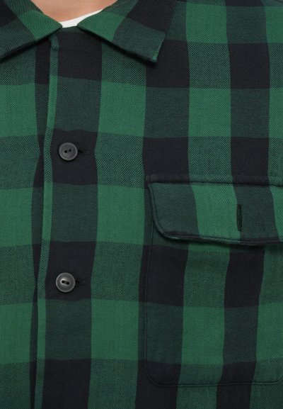 Camisa de flanela xadrez verde e preta, com colarinho, dois botões pretos e um bolso no peito com detalhe de aba com botão.