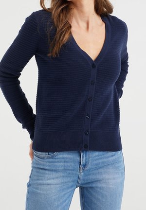 Femme portant un cardigan côtelé bleu marine à col en V boutonné et un jean bleu clair, les mains placées dans le dos.