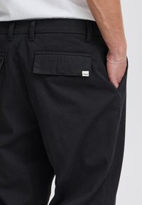 Svarte bukser med en jevn tekstur, med to baklommer og en liten hvit merkelapp. Linningen har beltestropper.