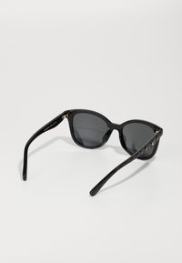 Lunettes de soleil noires en forme d'œil de chat, verres sombres, monture en plastique brillant, avec des accents dorés au niveau de la charnière. Détails texturés sur les côtés.